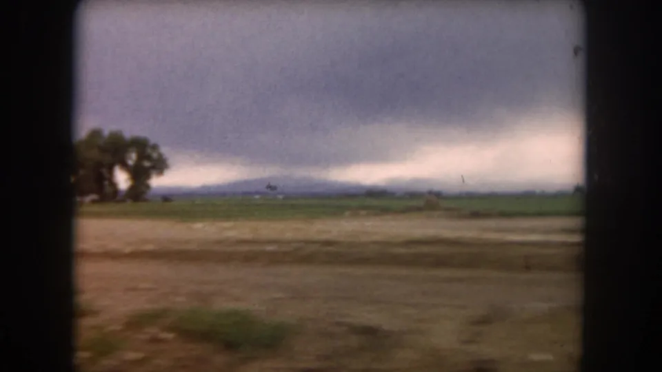1962 Summit County, Colorado rural field with a small tree cluster, distant low mountains, and a heavy cloudy sky.