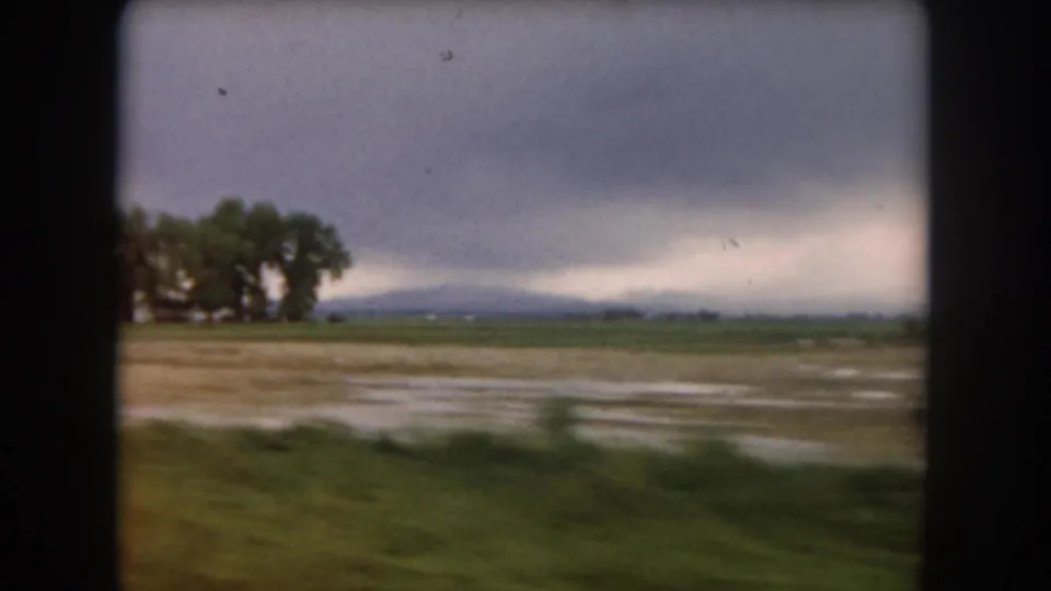 1962 Summit County, Colorado car view across a blurred grassy foreground, pale field, tree cluster, and distant mountains.