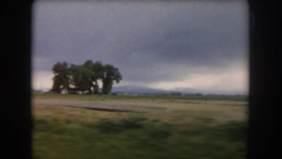 1962 Summit County, Colorado rural landscape with open field, roadside foreground blur, clustered trees, and distant mountains.