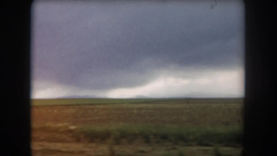 1962 Colorado home-movie frame showing open agricultural land and a low horizon beneath dense gray-white cloud cover.