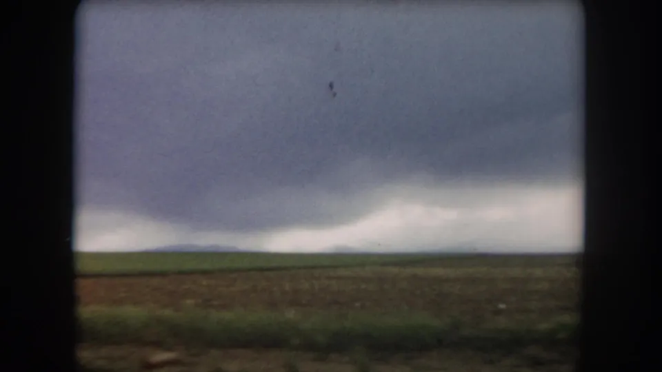 Soft 8mm frame of Summit County cultivated land, distant low hills, and layered clouds viewed from a moving vehicle.