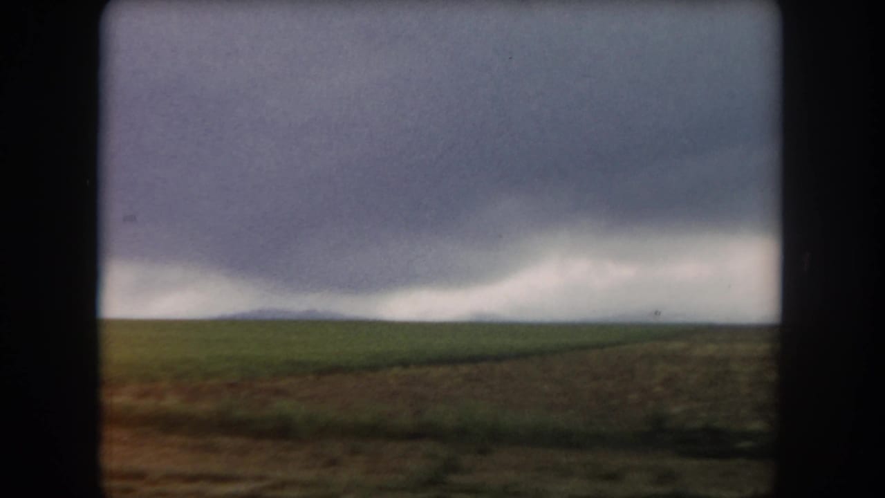 1962 Summit County, Colorado landscape frame with flat green ground, brown field edge, and a dark weather front beyond.