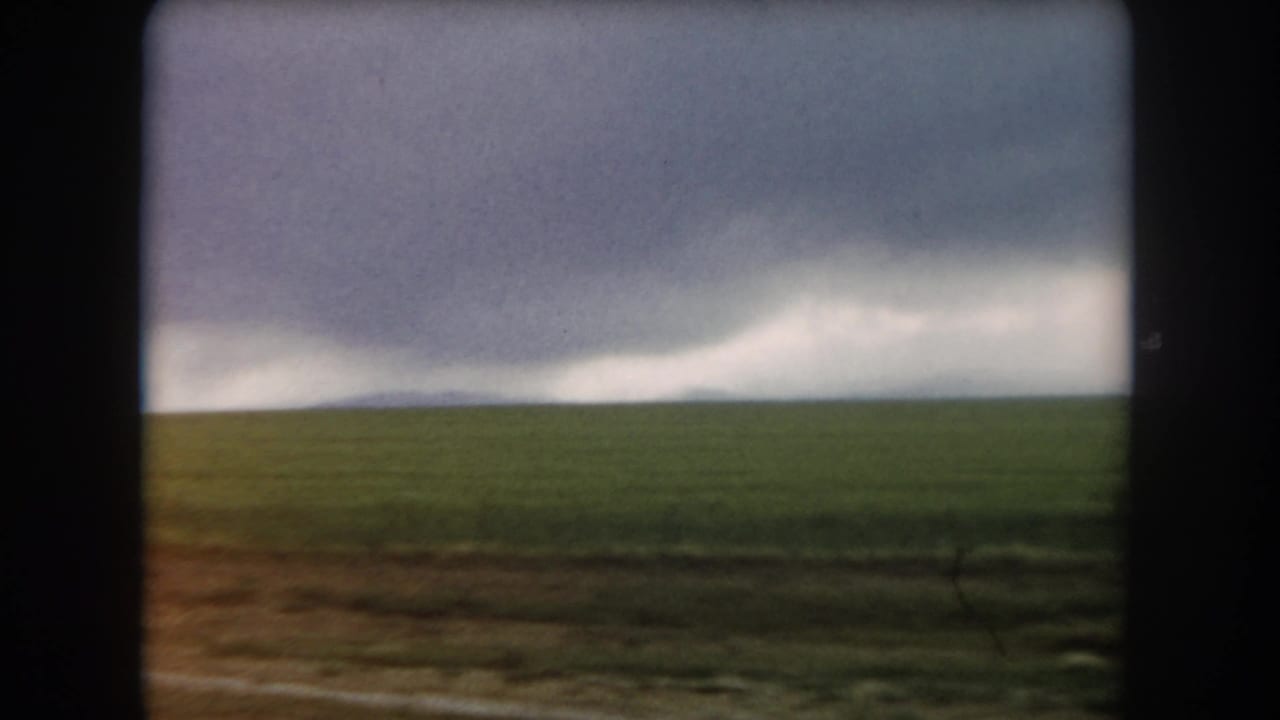 1962 Colorado home-movie view of open green field and brown roadside under a heavy, overcast sky.