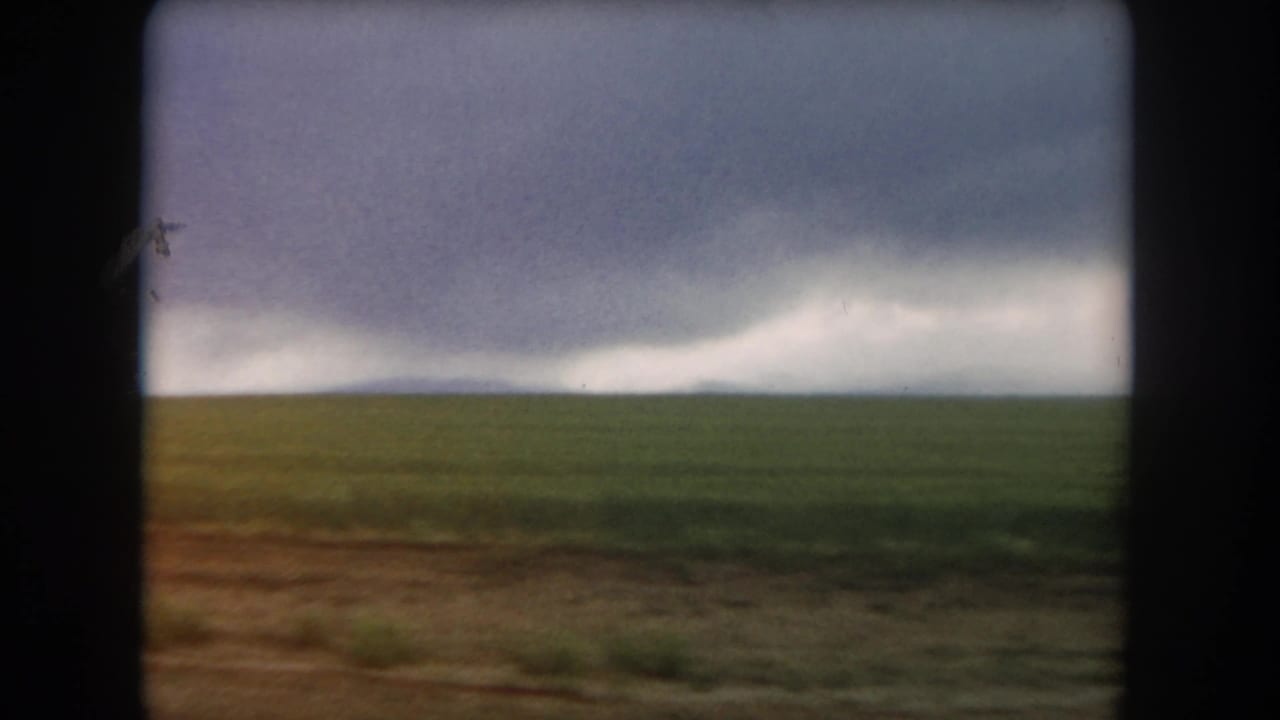 A moving 8mm view across Colorado plains with low green field, brown foreground, and storm clouds on the horizon.