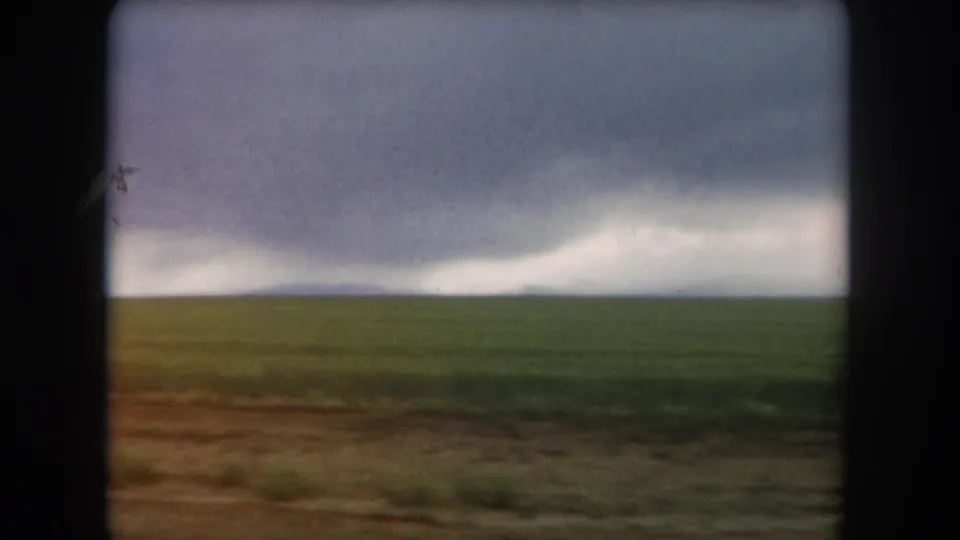 A moving 8mm view across Colorado plains with low green field, brown foreground, and storm clouds on the horizon.