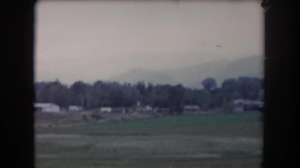 1962 Summit County view from a moving vehicle, showing fields, trees, low buildings, and hazy hills in the distance.