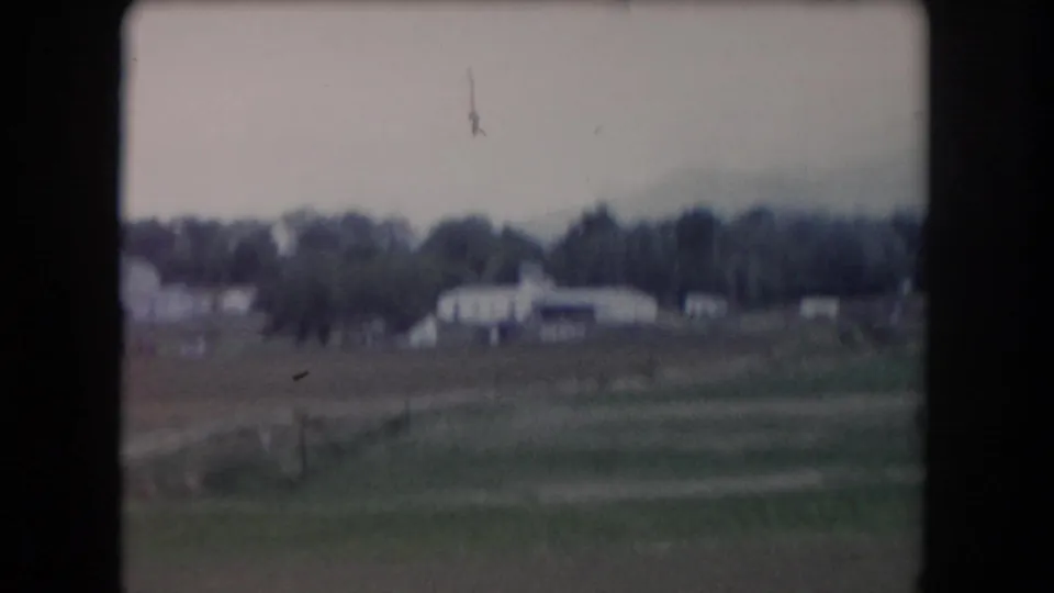 Summit County rural landscape on 8mm film, with open fields, pale farm buildings, trees, and a small dark object high in the cloudy sky.
