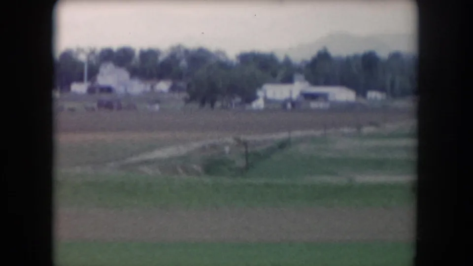 1962 8mm view of Summit County countryside, with distant pale buildings, a treeline, open fields, and a faint track or road across the ground.