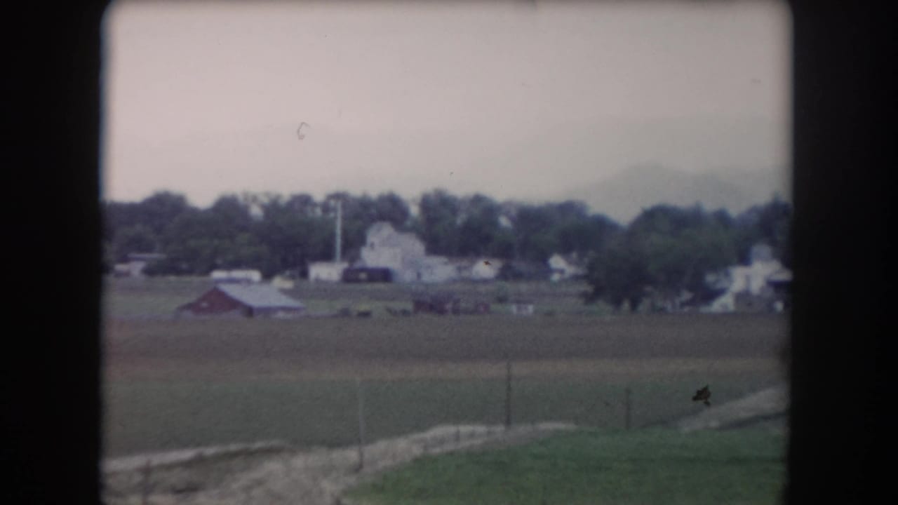 1962 Summit County farmland view with fence posts, red barn, white farm structures, trees, and mountains in haze.