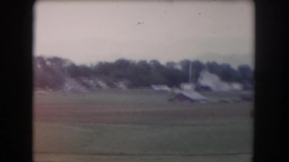 Summit County home-movie frame showing a distant farmstead beyond broad pasture under a hazy sky.