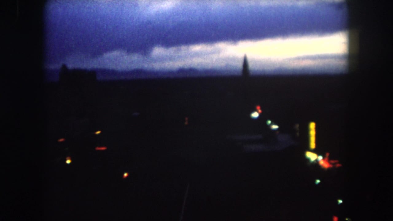 1962 8mm view of a dark Summit County, Colorado skyline at dawn, with a pale horizon, spire silhouette, tall building, and city lights below.