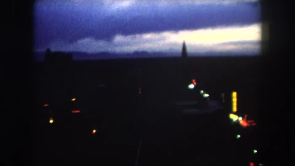 1962 8mm view of a dark Summit County, Colorado skyline at dawn, with a pale horizon, spire silhouette, tall building, and city lights below.