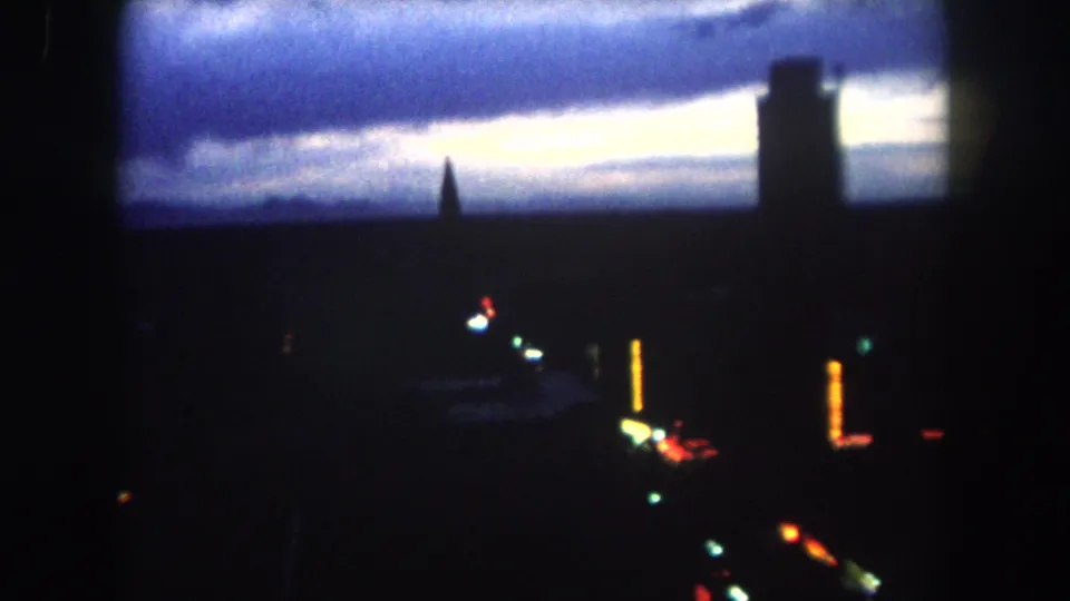 1962 8mm frame of a dawn skyline in Summit County, Colorado, with a central spire, tall dark building, pale sky, and blurred colored lights.