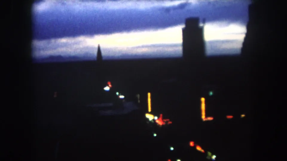 Dark 1962 8mm dawn view in Summit County, Colorado, with skyline silhouettes, a pointed spire, tall building mass, and scattered colored lights.