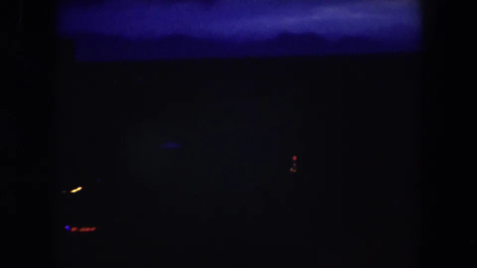 1962 8mm frame with a mostly black night landscape, faint urban silhouettes, and small red, yellow, and blue light points.