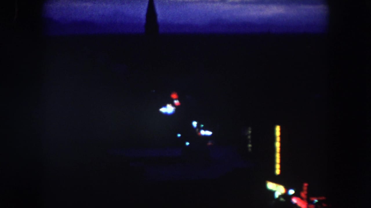 Dark aerial night view with colored lights, a spire silhouette, and bright vertical signage.