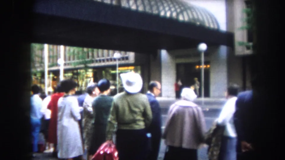 Adults in 1962 Colorado footage stand near a covered building entrance or awning, with a white hat and period coats visible.