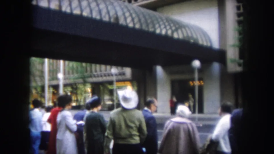 The 1962 Colorado home-movie frame shows adults in period clothing standing near a modern covered urban entrance.