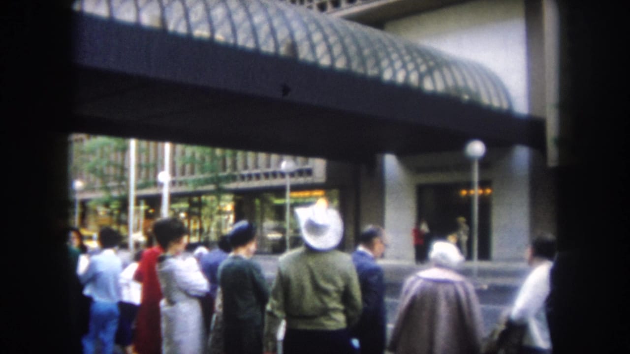 1962 home-movie frame of a sidewalk crowd waiting near a modern building entrance in Summit County, Colorado.