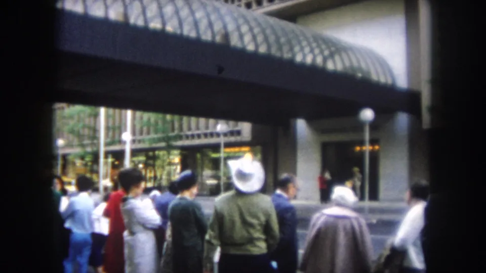 1962 home-movie frame of a sidewalk crowd waiting near a modern building entrance in Summit County, Colorado.