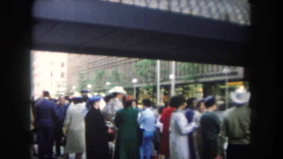 Handheld 1962 8mm view of people waiting on a sidewalk near tall buildings and a covered entrance.