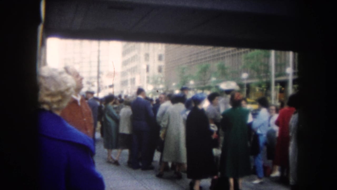 1962 Colorado-labeled 8mm frame showing adults gathered on a downtown sidewalk behind a woman in a blue coat, with tall buildings beyond.