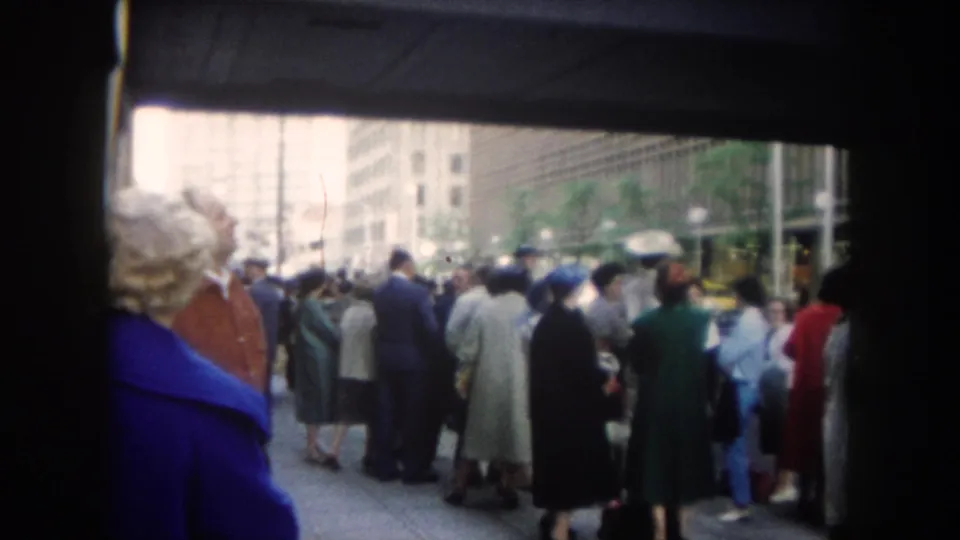 1962 Colorado-labeled 8mm frame showing adults gathered on a downtown sidewalk behind a woman in a blue coat, with tall buildings beyond.