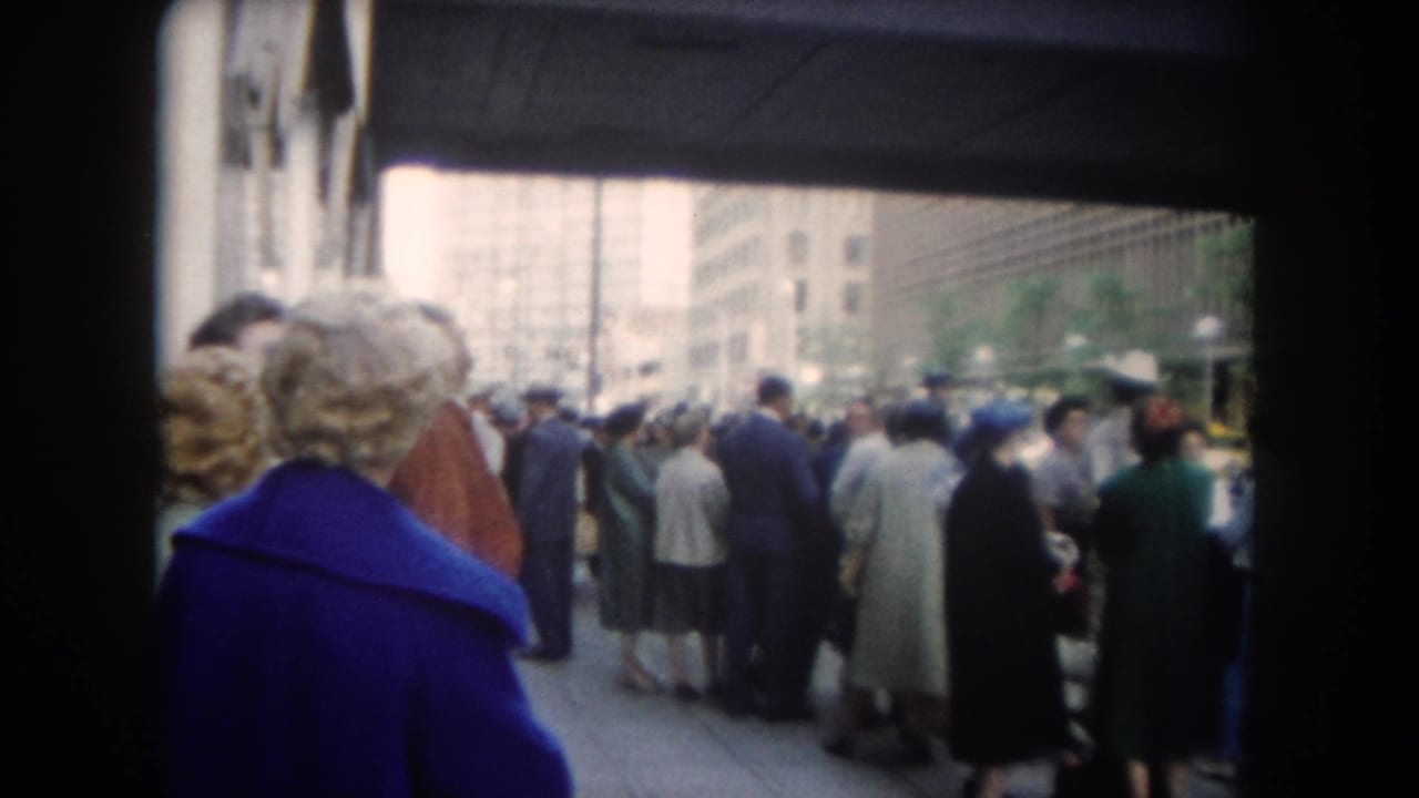 1962 Colorado-labeled home-movie frame of a city sidewalk crowd in coats and hats, filmed through a dark surrounding frame.