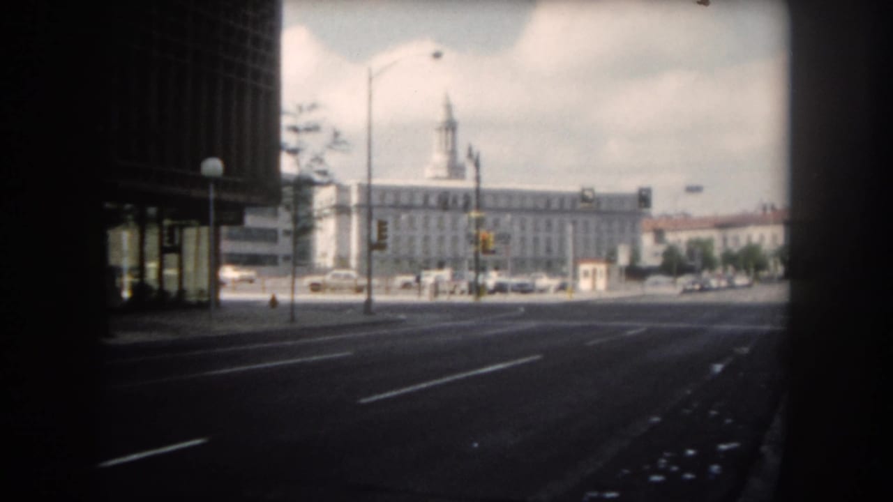 1962 8mm home-movie view of a quiet street in Summit County, Colorado, framed by shadow with parked cars and a towered building beyond.