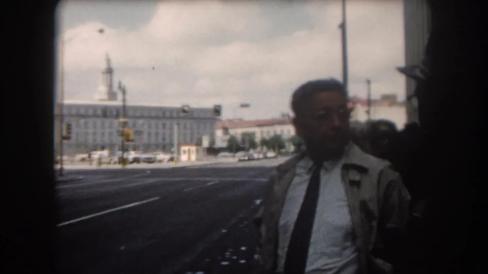 Adult man close to the camera in a 1962 8mm Summit County street view, with the broad road and distant towered building behind him.