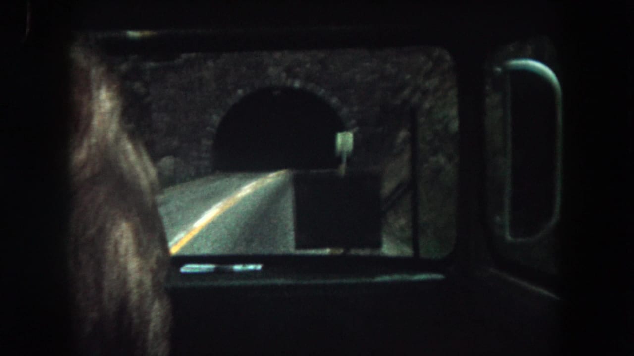 Bus window view of a road leading toward a dark stone tunnel.