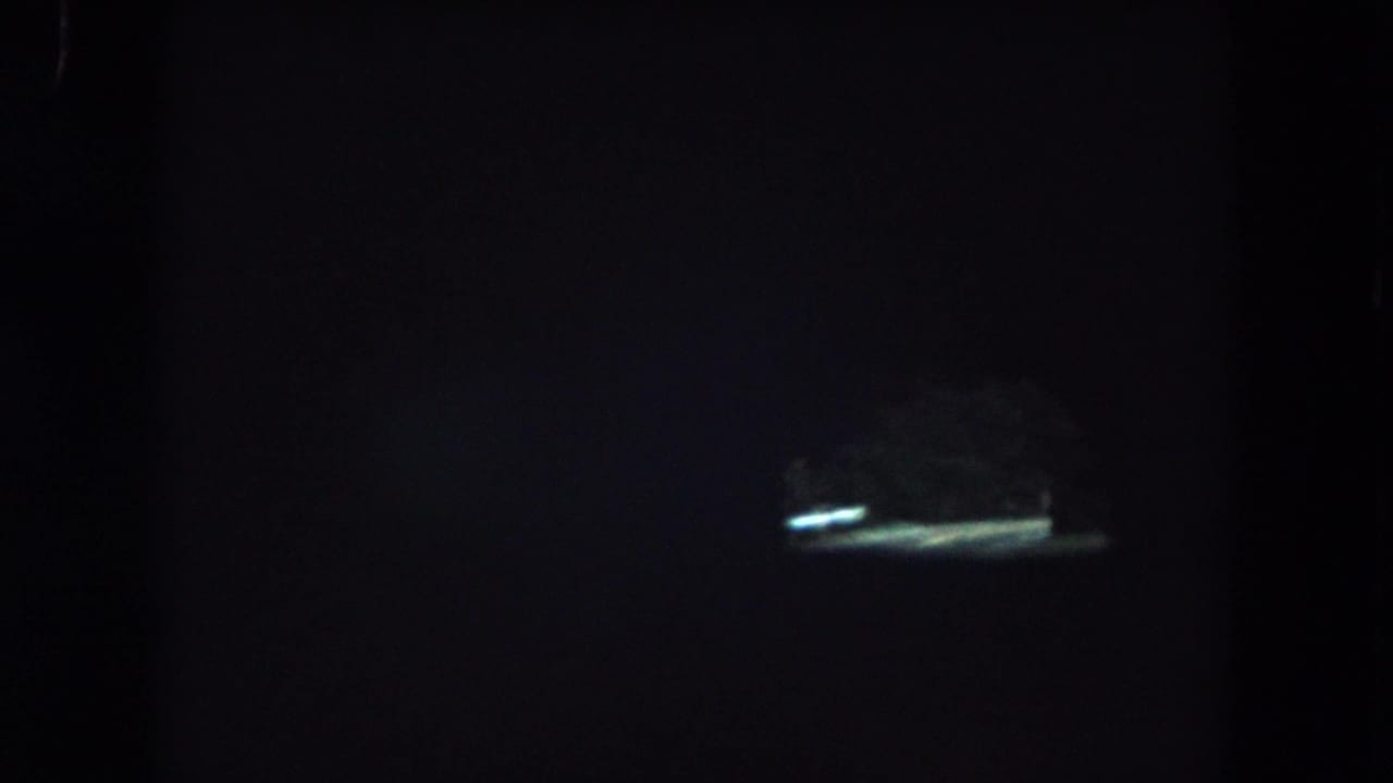 Low-light 8mm bus interior view, with the windshield framing a bright opening or reflected light ahead in the tunnel.