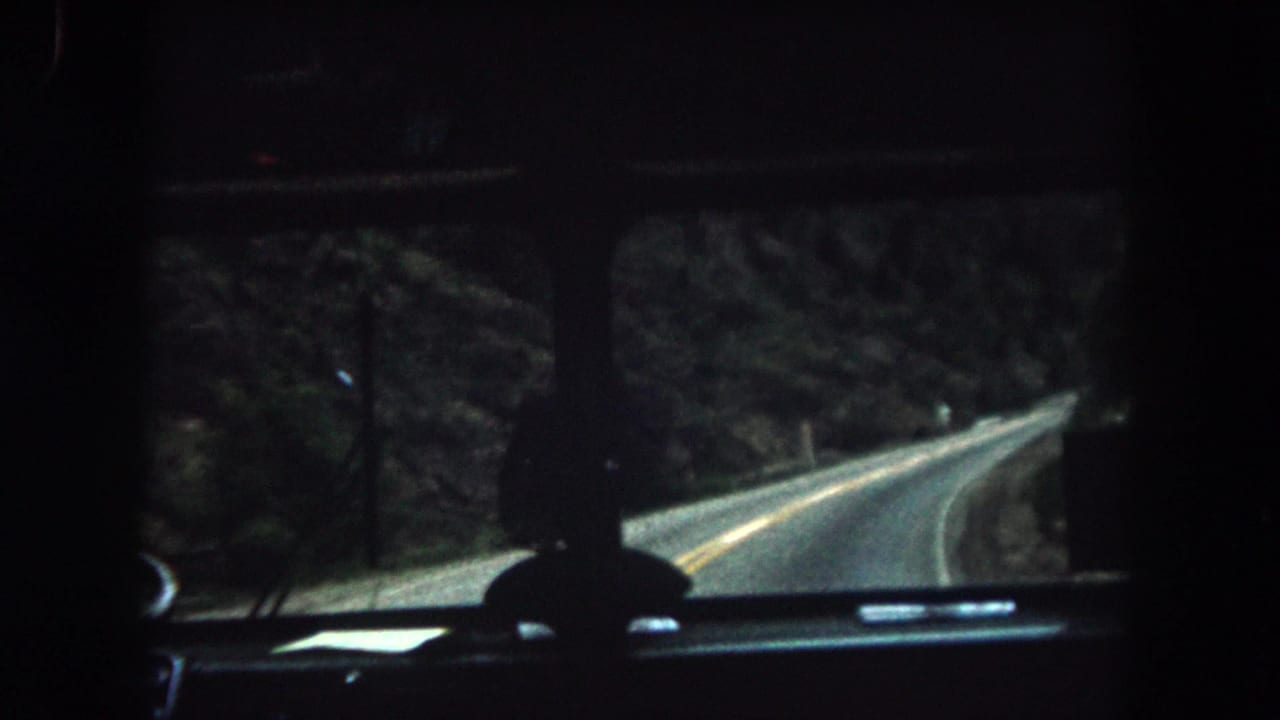 1962 Summit County, Colorado home-movie frame showing a dim vehicle interior and winding two-lane road through forest.