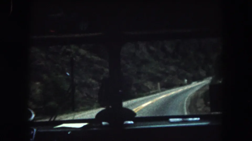 1962 Summit County, Colorado home-movie frame showing a dim vehicle interior and winding two-lane road through forest.