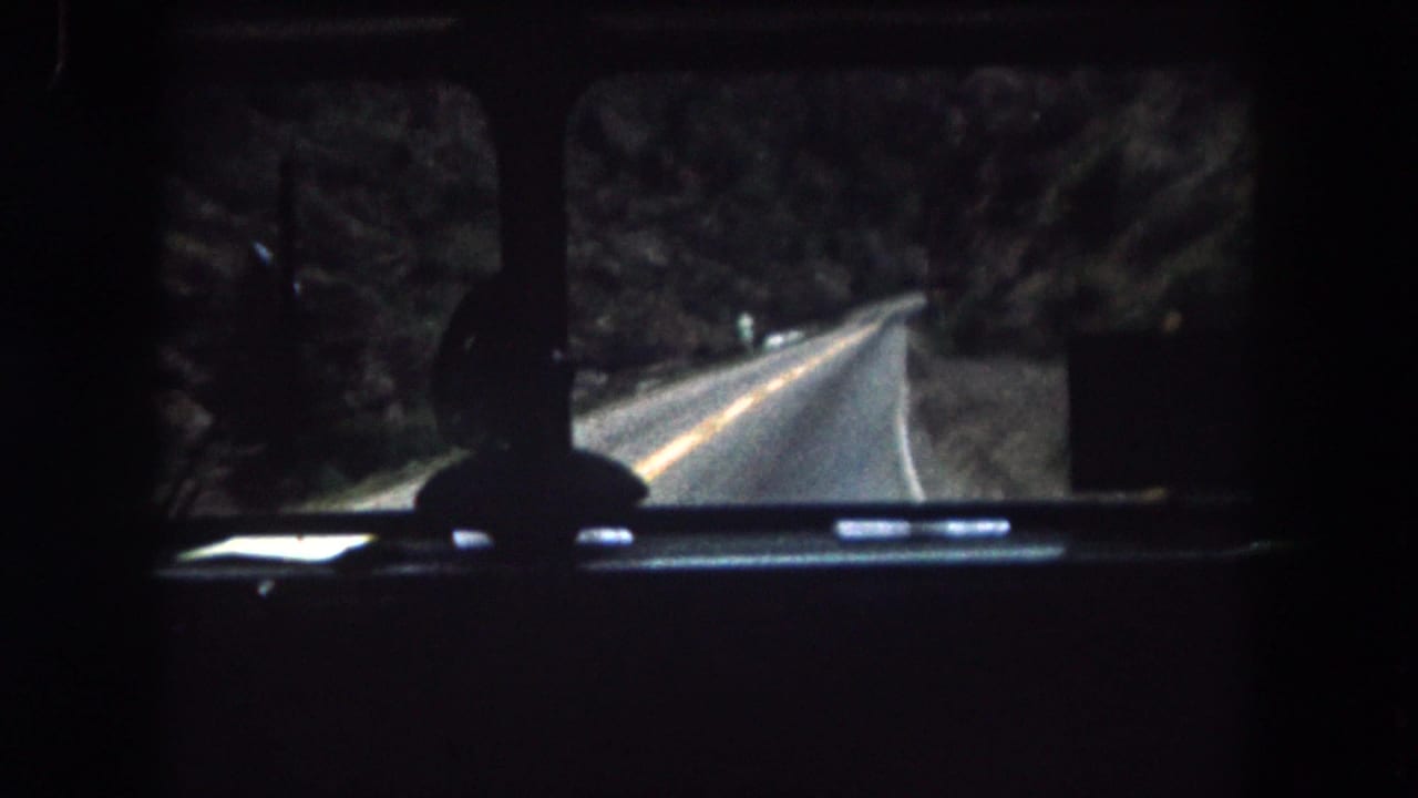 Windshield view from a 1962 Colorado vehicle interior, with a narrow road bending through trees and rocky terrain.