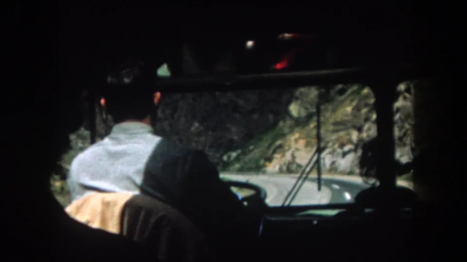 A man drives toward a bend in a rocky mountain road, filmed from the rear seat in 1962 Colorado footage.