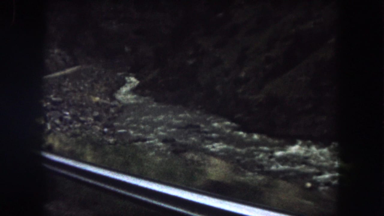 1962 Summit County, Colorado home-movie frame looking from a moving road past a guard rail toward a shadowed river or creek.