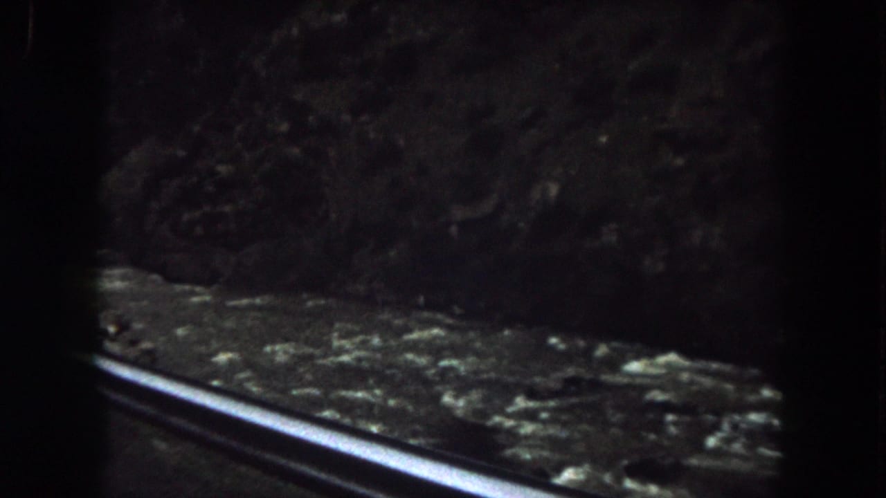 1962 Summit County, Colorado 8mm frame showing a shadowed rocky embankment and roadside guardrail from a moving vehicle.
