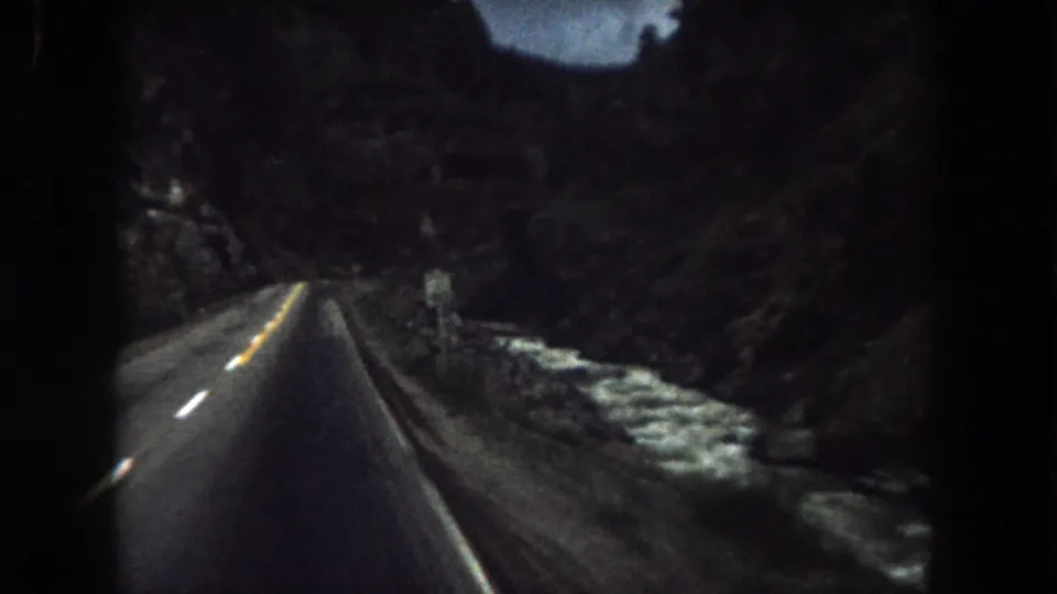 1962 Summit County, Colorado frame looking down a paved mountain road with a guardrail, cliff walls, and pale roadside patches.
