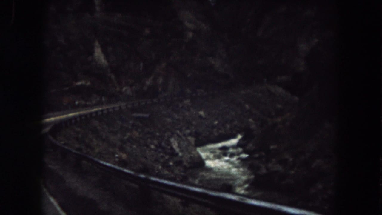 1962 Colorado train-window view with a curved rail line, rocky embankment, and bright stream water in a canyon.