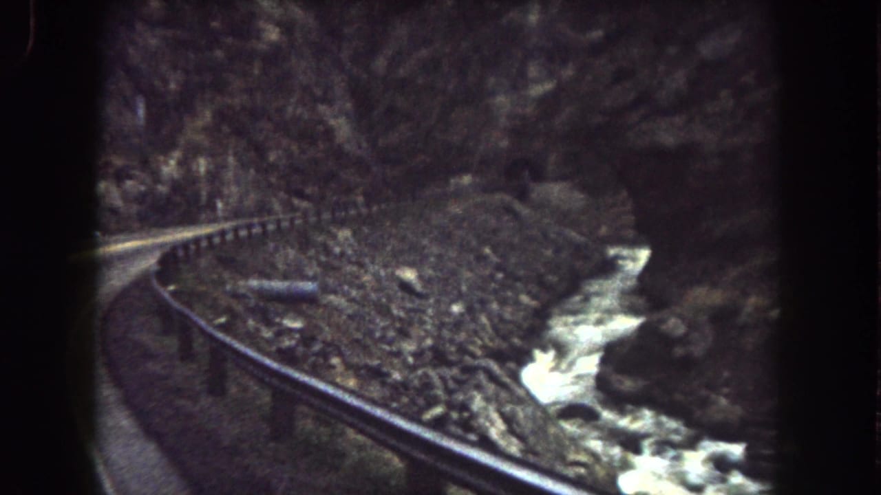 1962 Summit County, Colorado frame with a canyon road, roadside guardrail, rocky embankment, and white water in the stream.