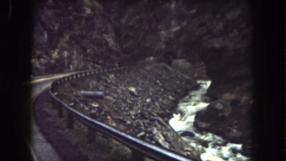 1962 Summit County, Colorado frame with a canyon road, roadside guardrail, rocky embankment, and white water in the stream.