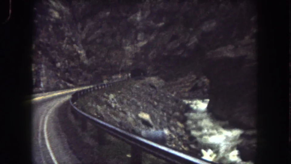 1962 Summit County, Colorado 8mm frame of a winding road, guardrail, canyon rocks, and bright stream water.