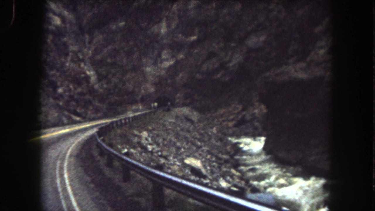 1962 Summit County home-movie view of a car approaching a bend on a two-lane mountain road near a rocky canyon and guardrail.