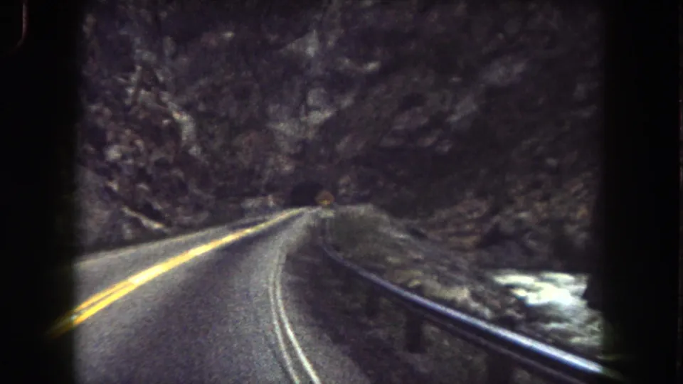 Vehicle-level 8mm view of a paved two-lane road through rocky Summit County terrain, with guardrail and roadside water or snow.