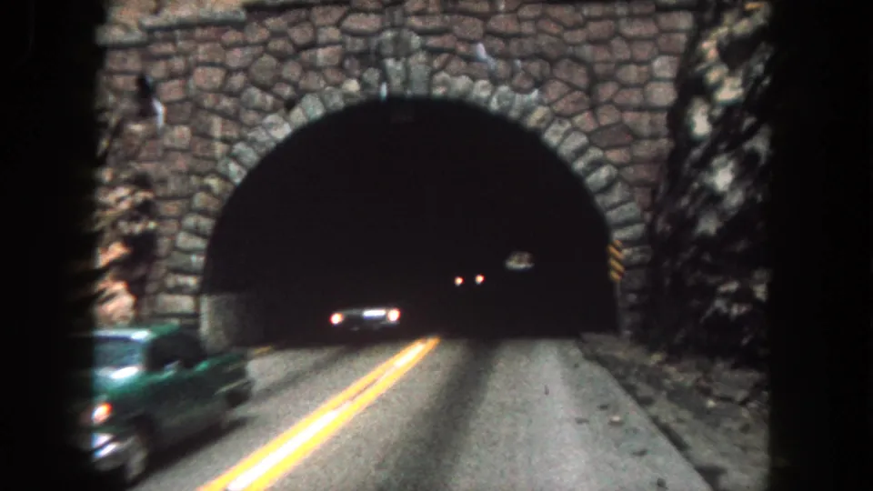 A green car passes in the foreground while another vehicle is visible near the dark tunnel entrance on the mountain road.