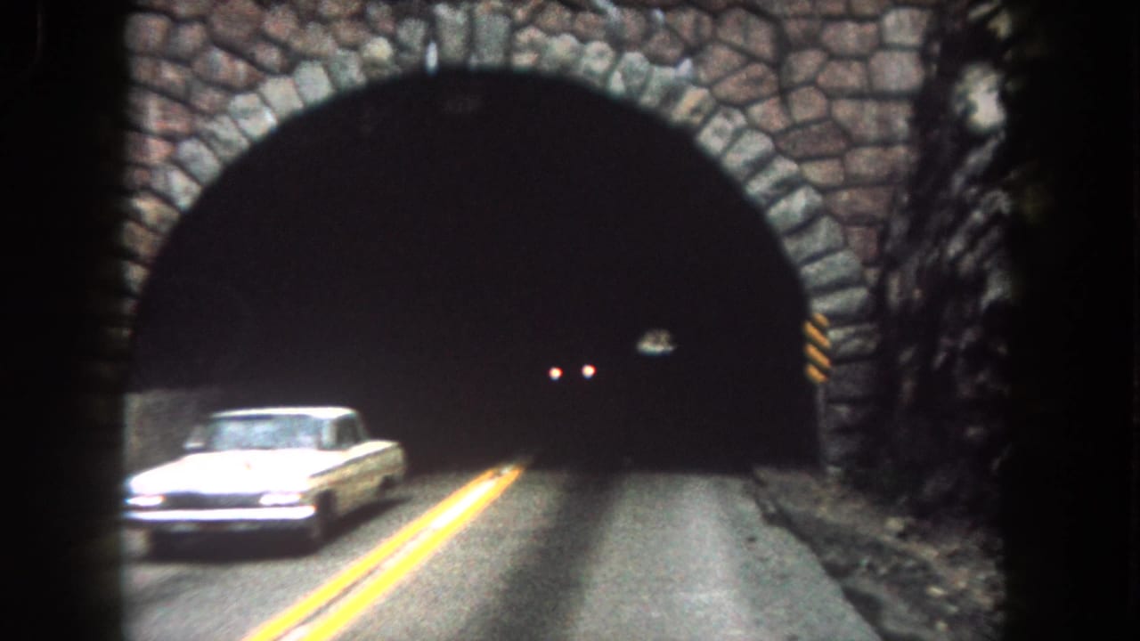 1962 Summit County home-movie frame showing a white car near a stone-lined tunnel entrance with yellow lane lines.