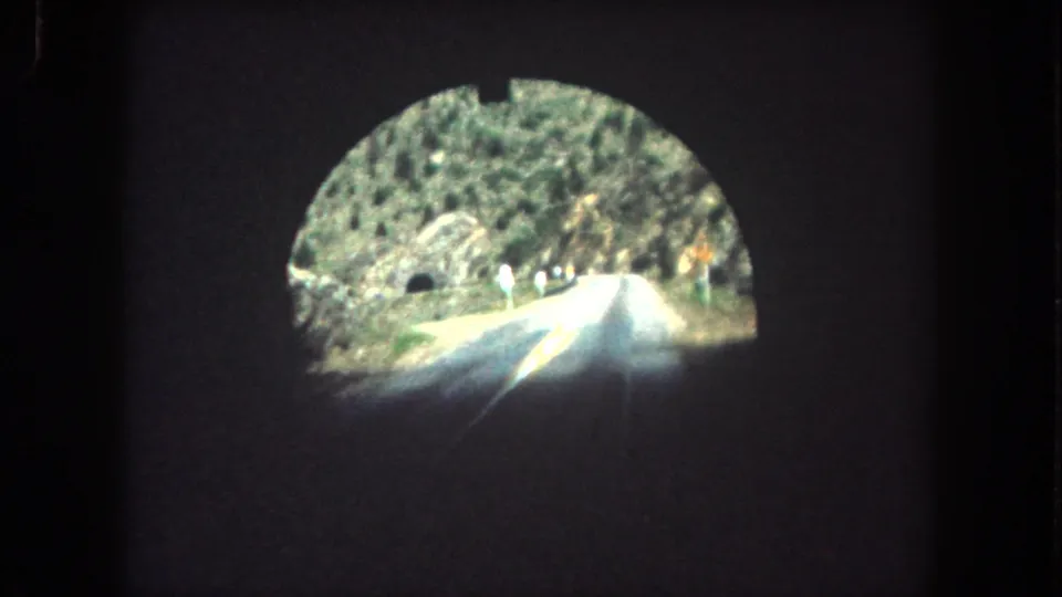 1962 8mm view emerging from a Summit County tunnel onto a narrow mountain road with a yellow center line and rocky slopes.