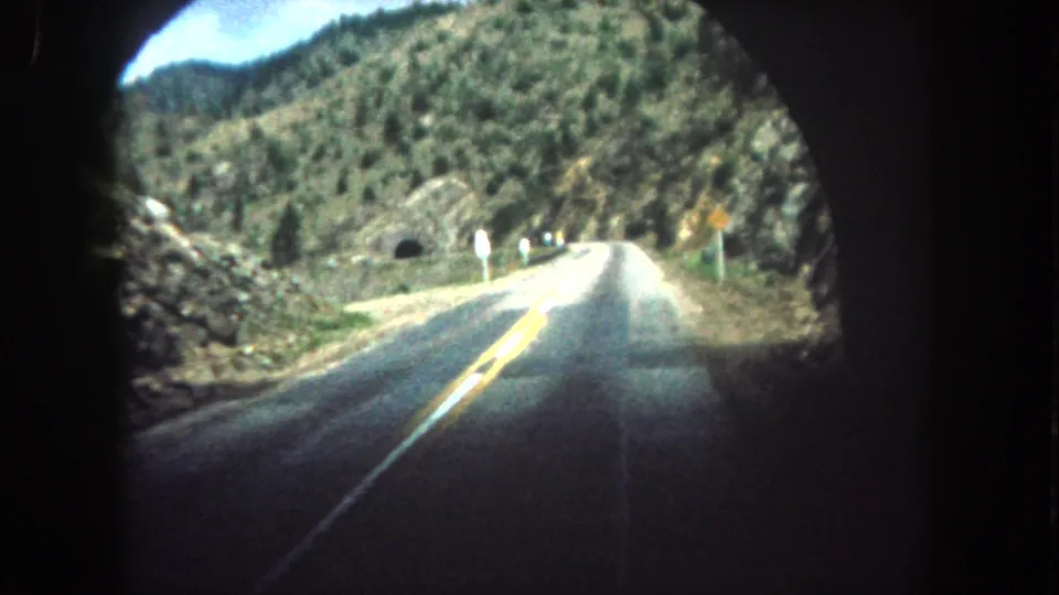Vehicle POV on a Summit County mountain road after leaving the tunnel, with rocky slopes, evergreen trees, and bright 8mm exposure.