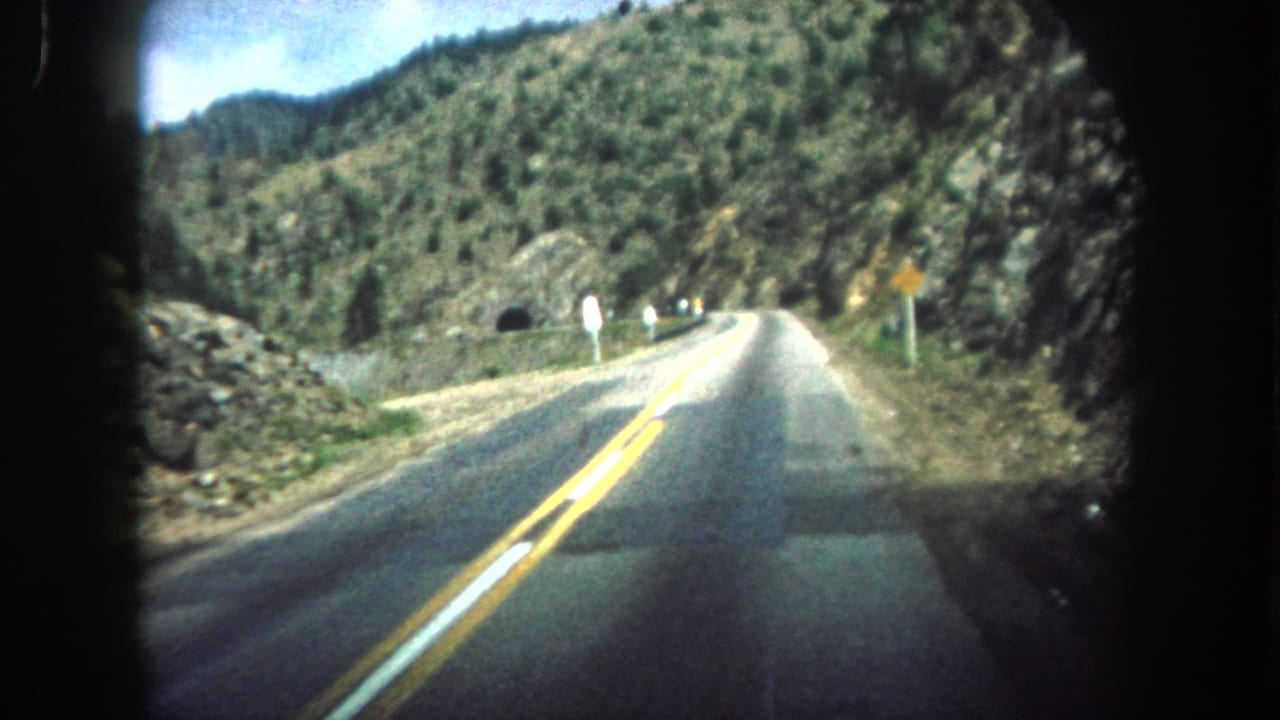 Vehicle POV on a two-lane mountain road with yellow center lines and rocky hillsides.